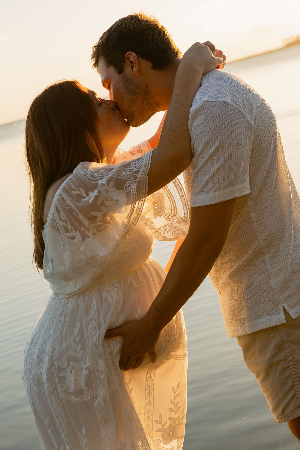 Couple sharing a romantic kiss at sunset by the water, woman in white lace dress, golden hour glow.