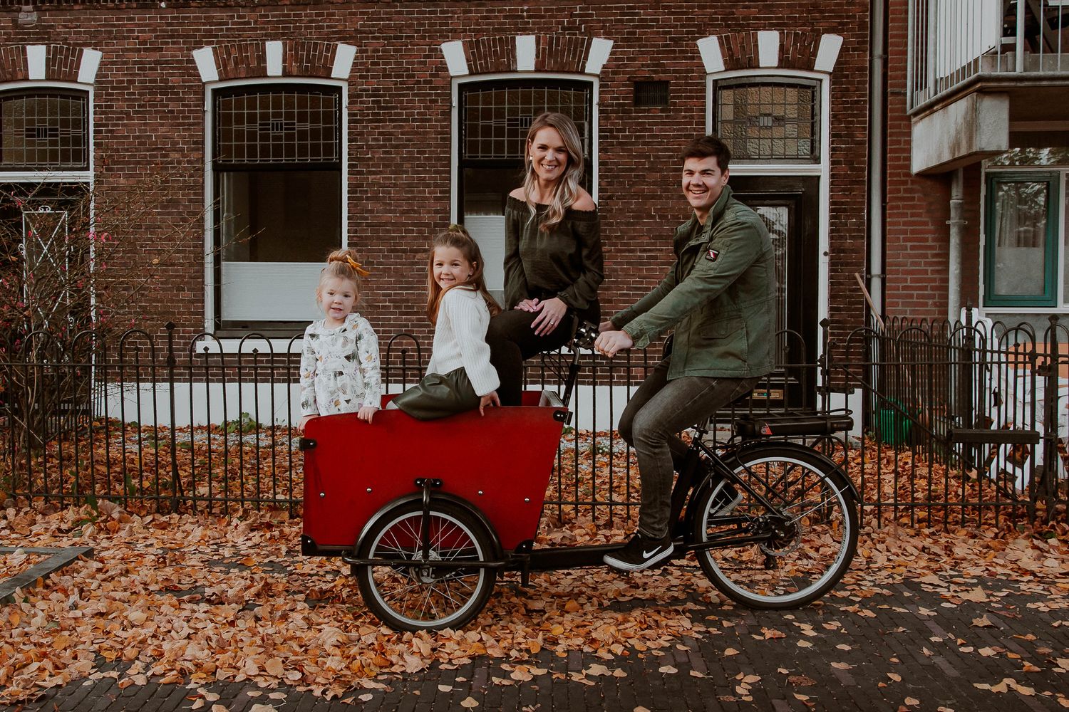 Family of four posing with a red cargo bakfiets on a leaf-covered street in autumn, in front of a brick building in Woerden.