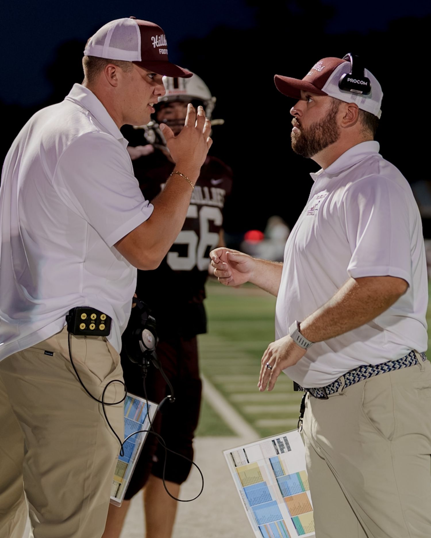 Two football coaches in white polos communicating on the sideline during a night game.