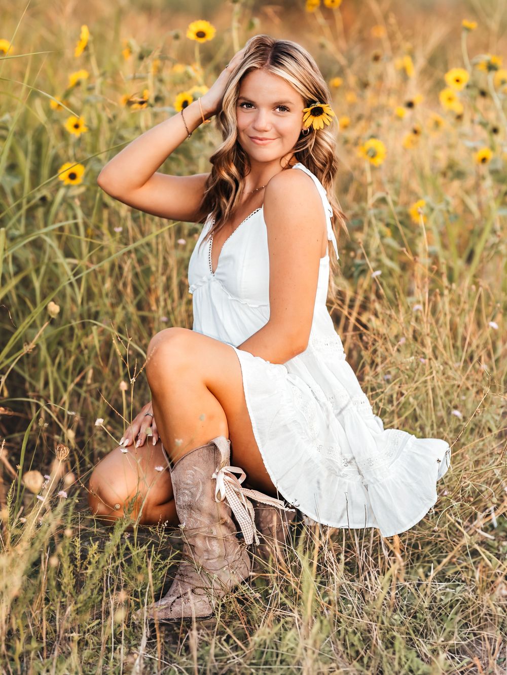 Young woman in white dress and cowboy boots poses in a sunflower field for a senior portrait photo.