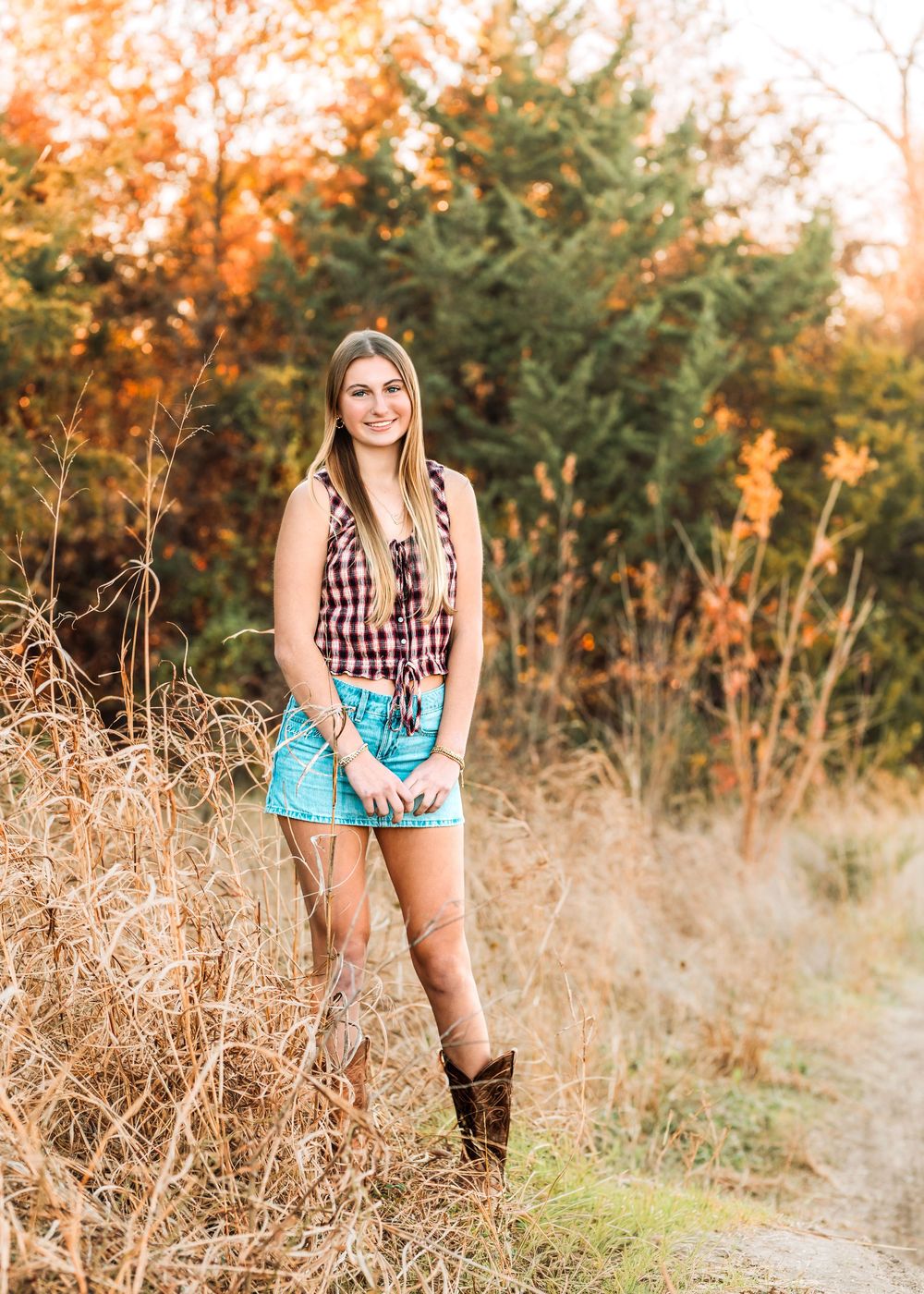 Young woman in plaid top, denim shorts, and brown boots poses outdoors among autumn foliage and dry grass.