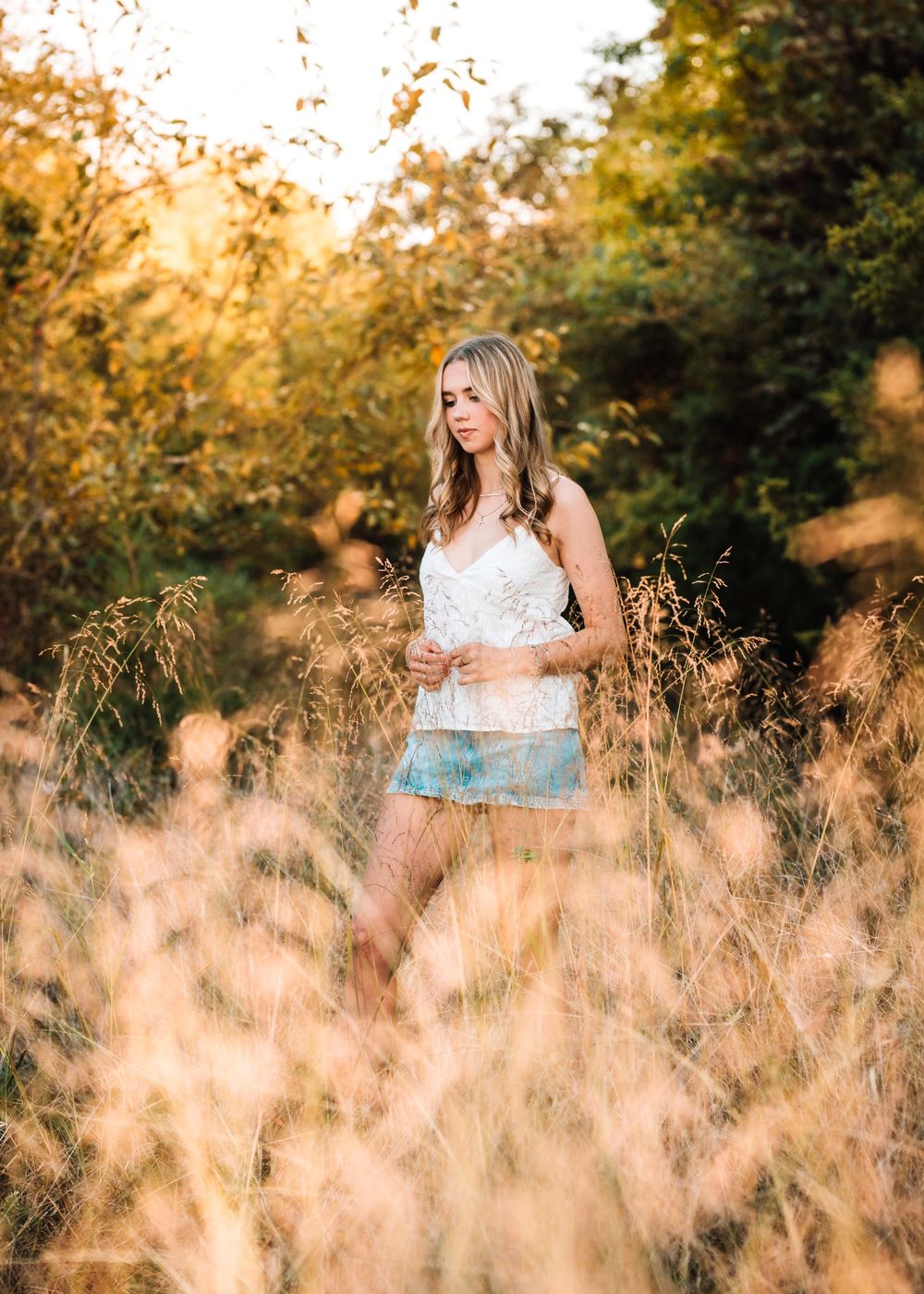 Young blonde woman in white top and denim shorts standing in golden tall grass field during sunset.