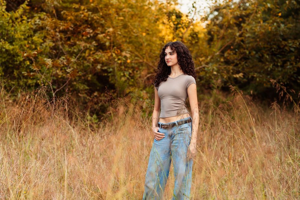 Young woman with curly hair in beige crop top and wide-leg jeans standing in tall grass with autumn trees.