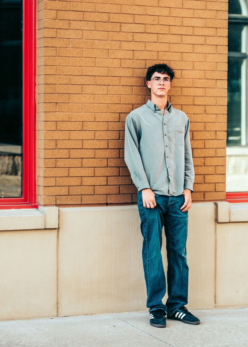 A young man in a gray button-up shirt and jeans leans against a brick wall outdoors.