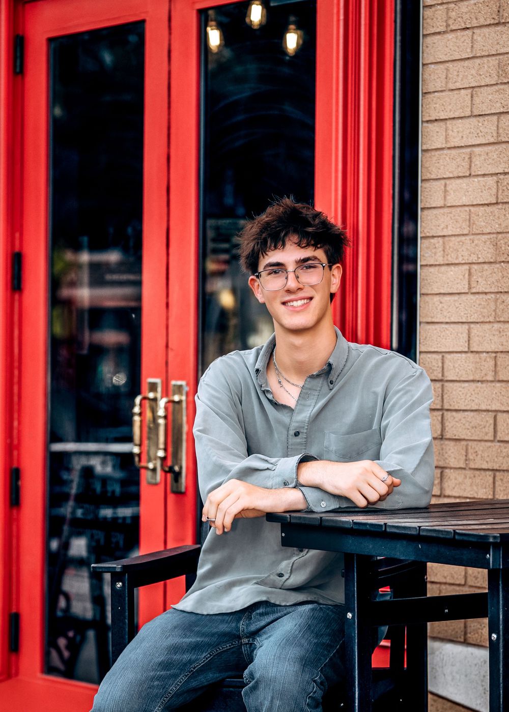 Young man with curly hair and glasses smiling while sitting at an outdoor table near red doors.