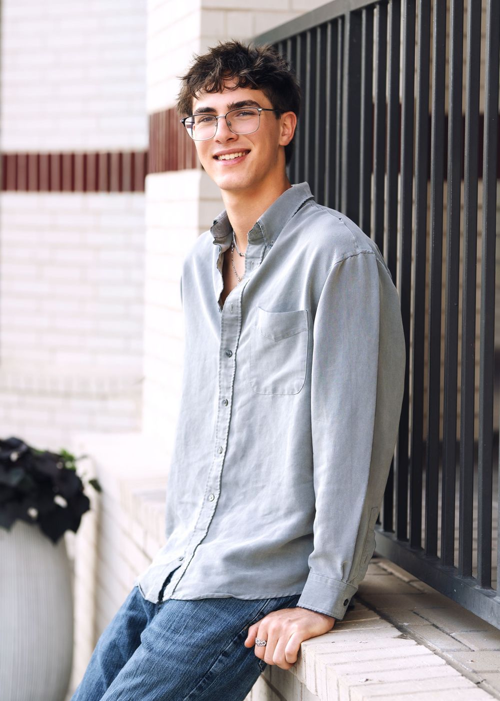 Smiling young man with curly hair and glasses wearing a light blue button-up shirt leaning against a wall outdoors.