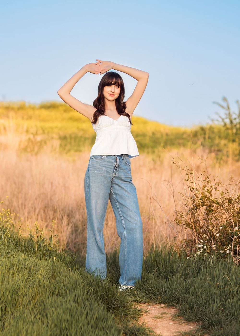 Young woman in white top and wide-leg jeans poses with arms raised in a sunny field.