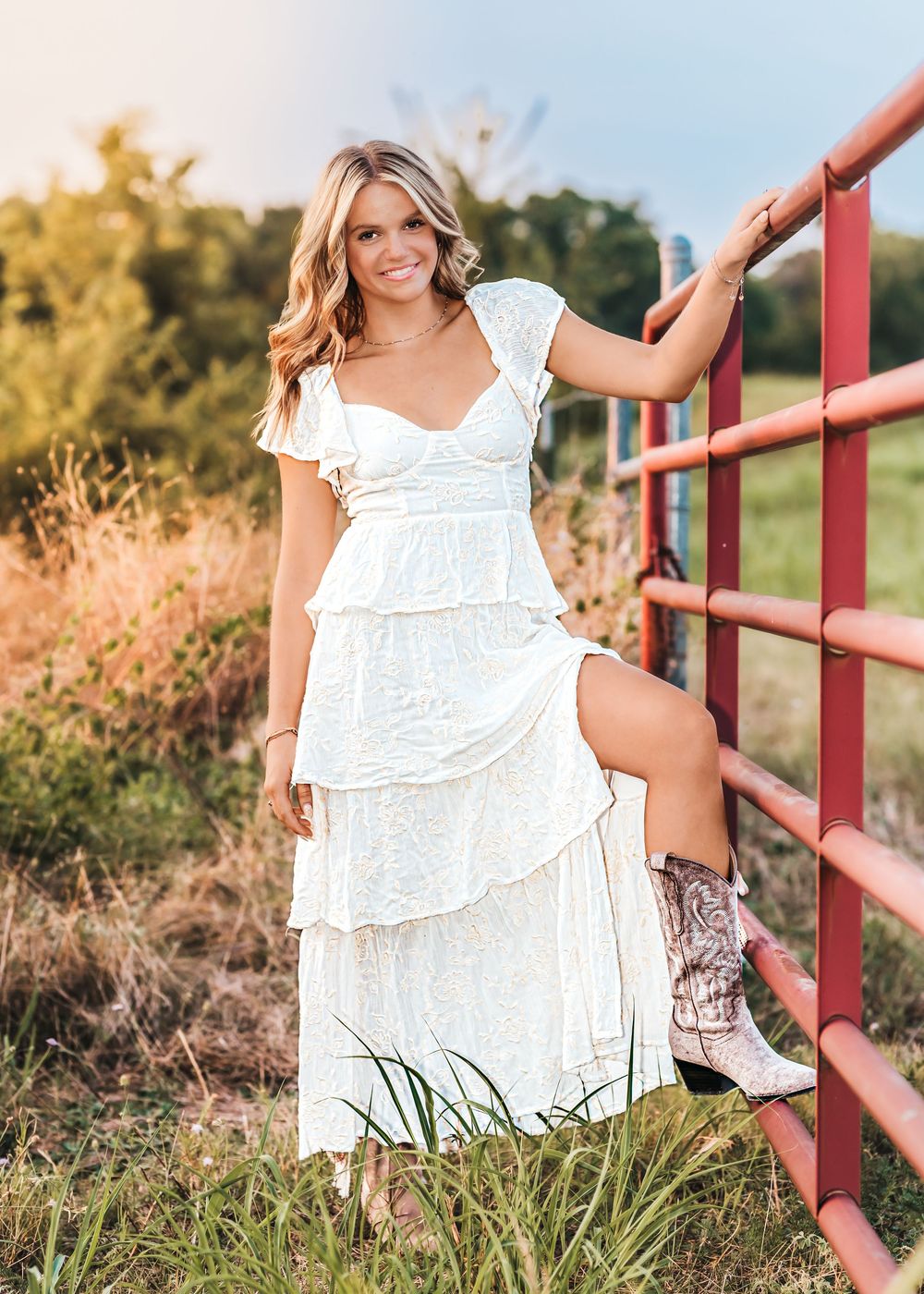 Young woman in white lace tiered maxi dress and cowboy boots leaning on red farm fence at golden hour.