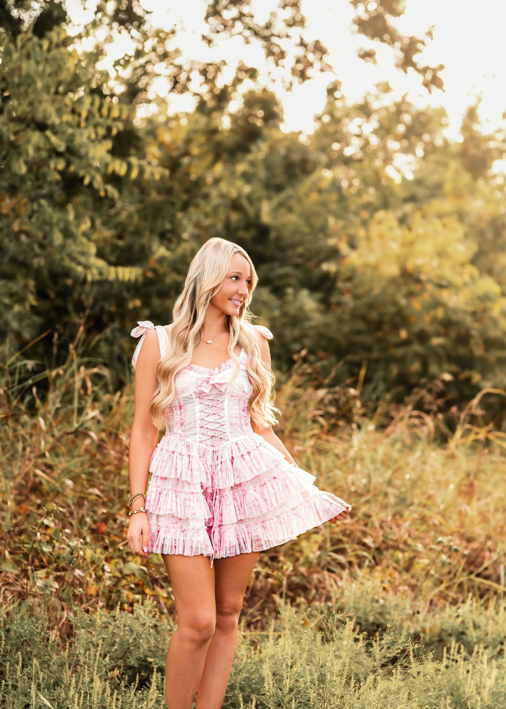 Young woman in pink ruffled mini dress posing outdoors in golden hour light with lush green trees behind her.