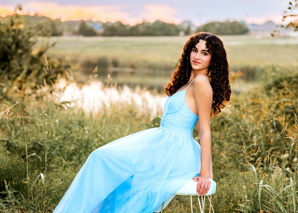 Young woman in a light blue gown poses gracefully in a lush green field near a pond at sunset.