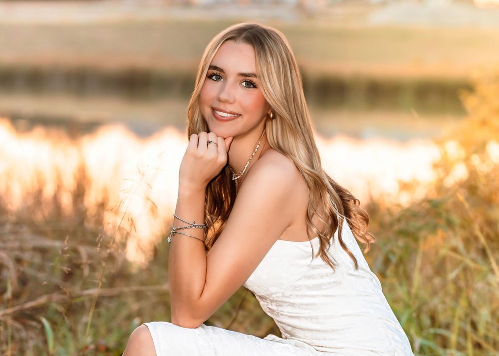 Young blonde woman in white dress smiling outdoors near water during golden hour sunset photography.