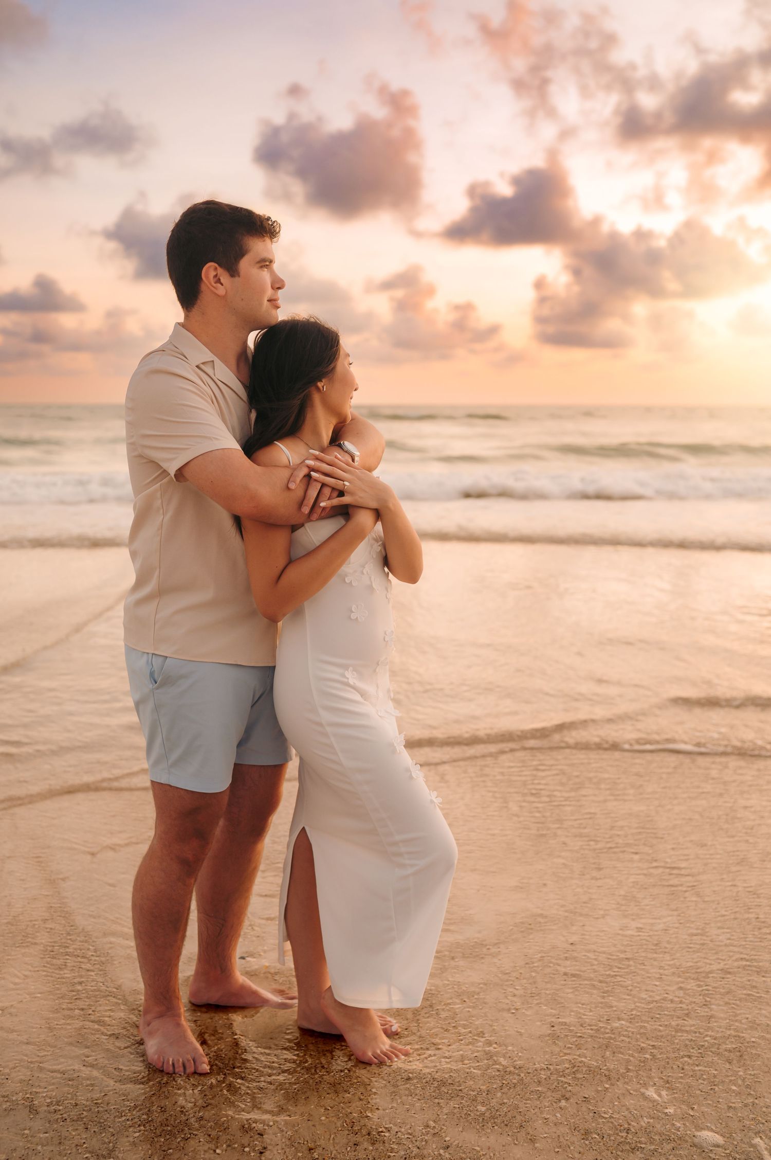 Couple embracing on a beach at sunset, standing barefoot on wet sand with colorful clouds in the background.