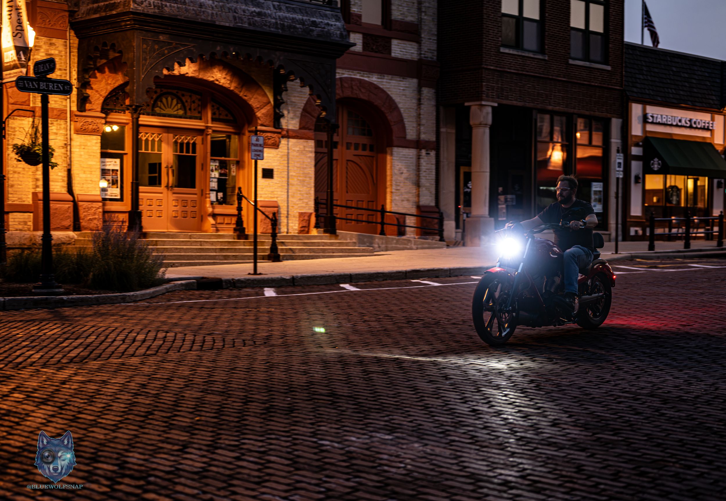 Motorcycle with bright headlight on cobblestone street at night near historic brick buildings.