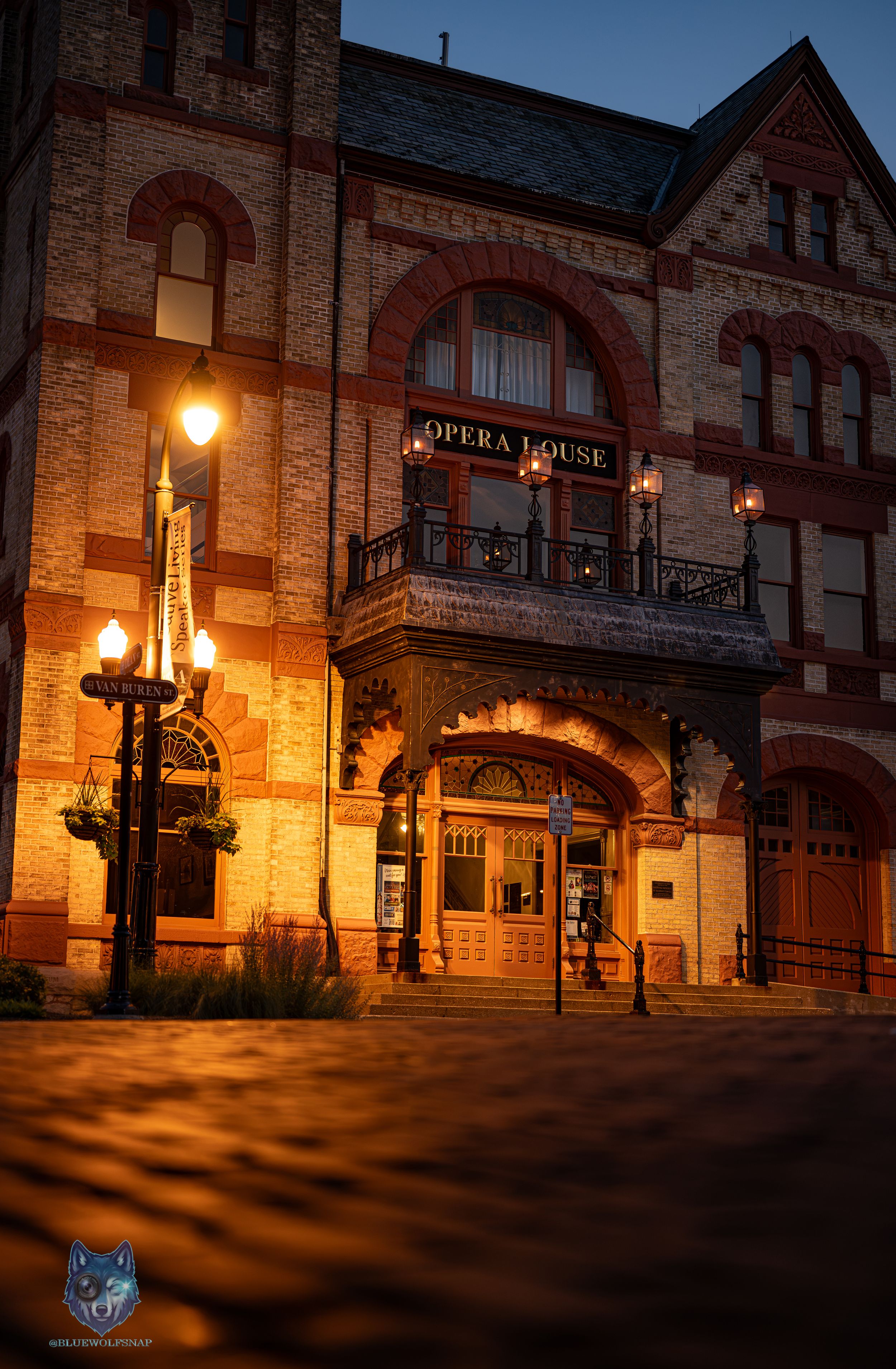 Historic Opera House building at dusk, illuminated by warm street lamps, featuring ornate red brick Victorian architecture.