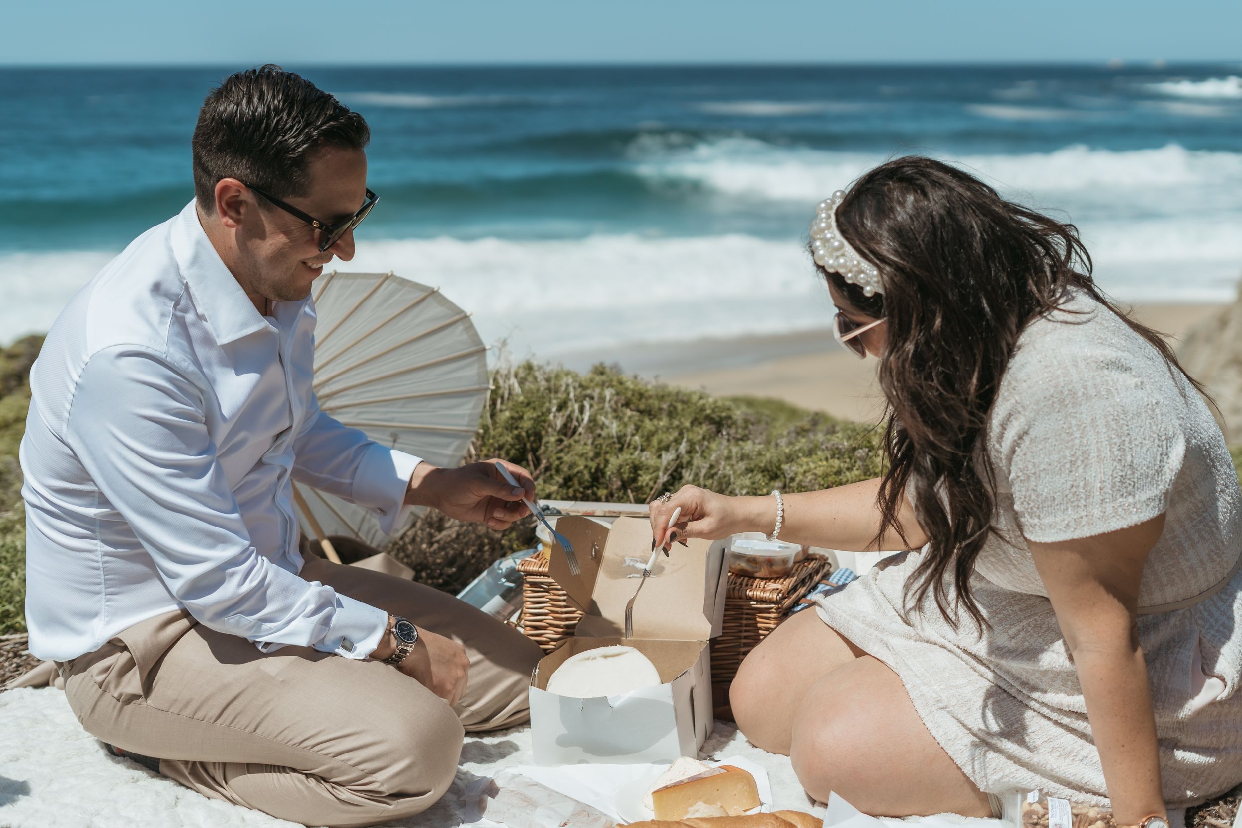 Couple enjoying a romantic beach picnic, sharing food on a white blanket with ocean waves in the background.