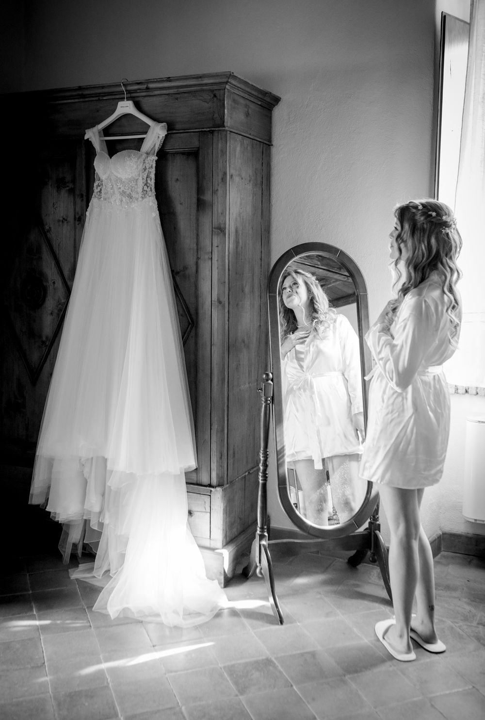 Bride in white robe admires wedding gown hanging on wardrobe beside floor mirror in elegant black and white photo.