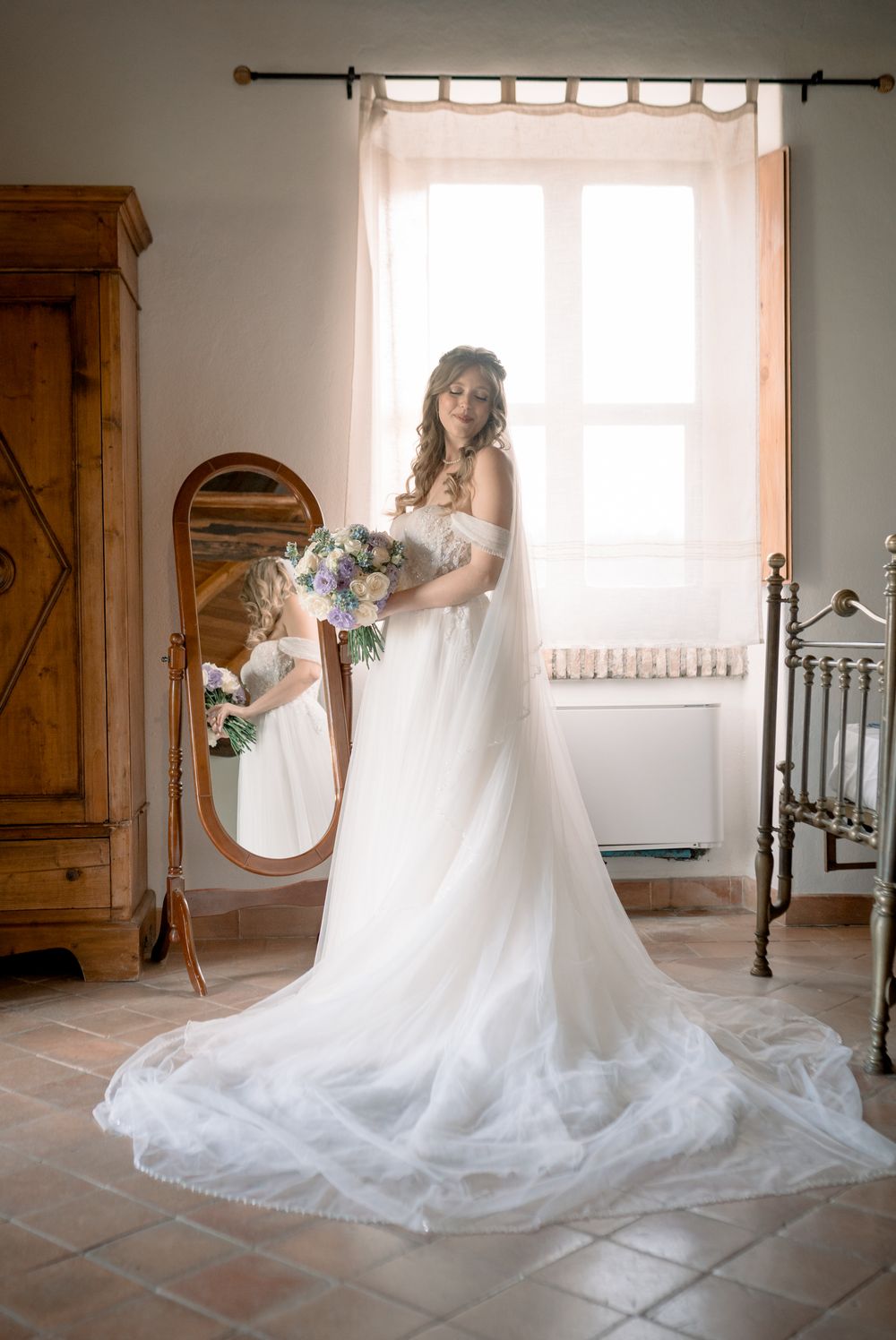 Bride in white off-shoulder gown with long train holds bouquet by oval mirror and window in rustic room.