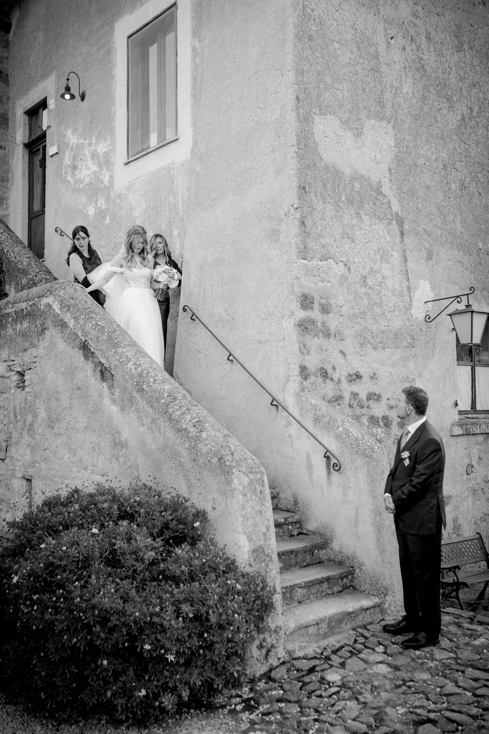 Black and white wedding photo of bride descending stone stairs toward groom outside rustic European building.