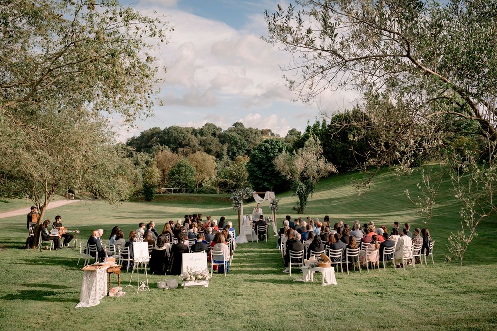 Outdoor wedding ceremony on lush green lawn with guests seated in rows facing floral arch under sunny sky.
