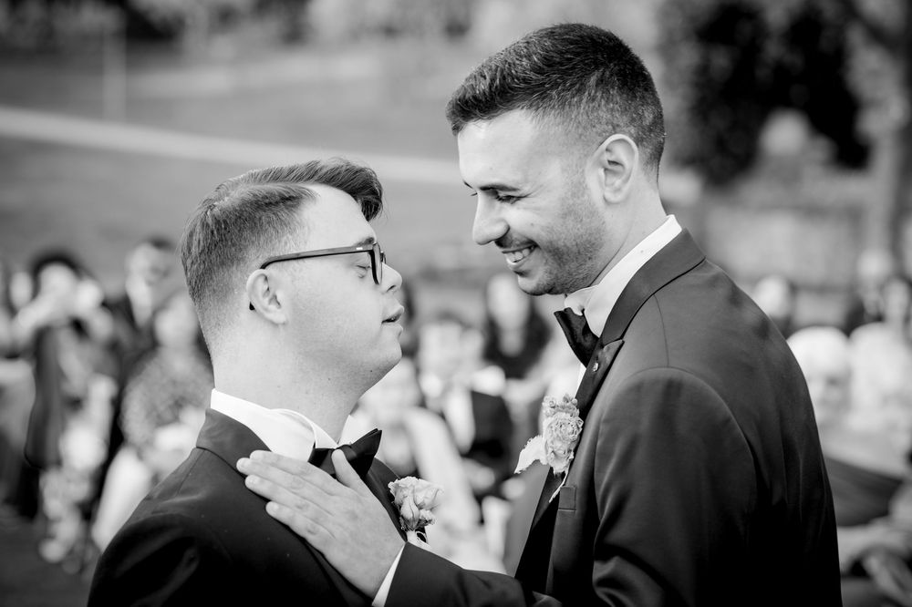 Two grooms share a tender moment at their wedding ceremony, dressed in elegant black tuxedos with bow ties.