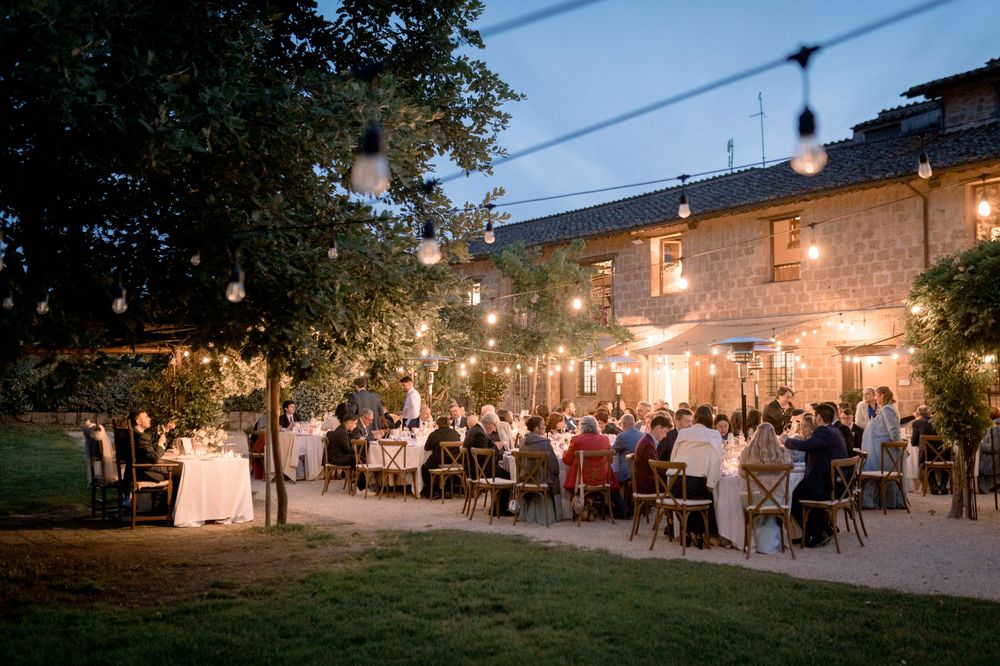 Outdoor wedding reception at dusk with string lights, guests dining at tables beside a rustic stone venue.