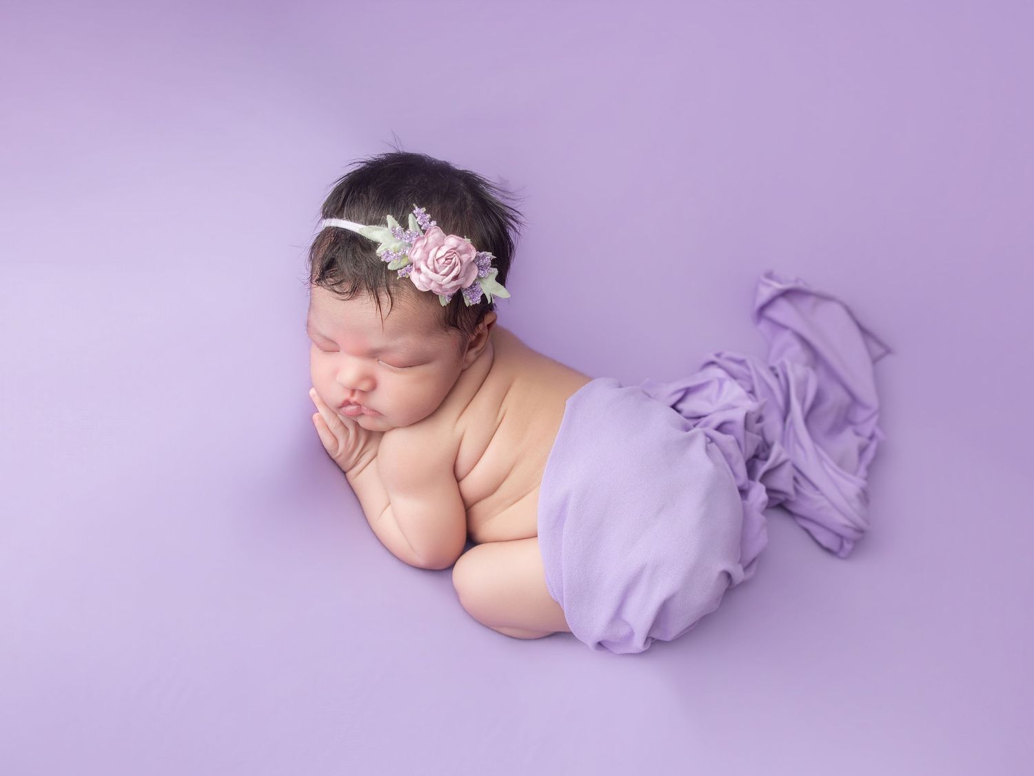 Newborn baby girl sleeping peacefully on purple backdrop, wrapped in lavender fabric with a floral headband.