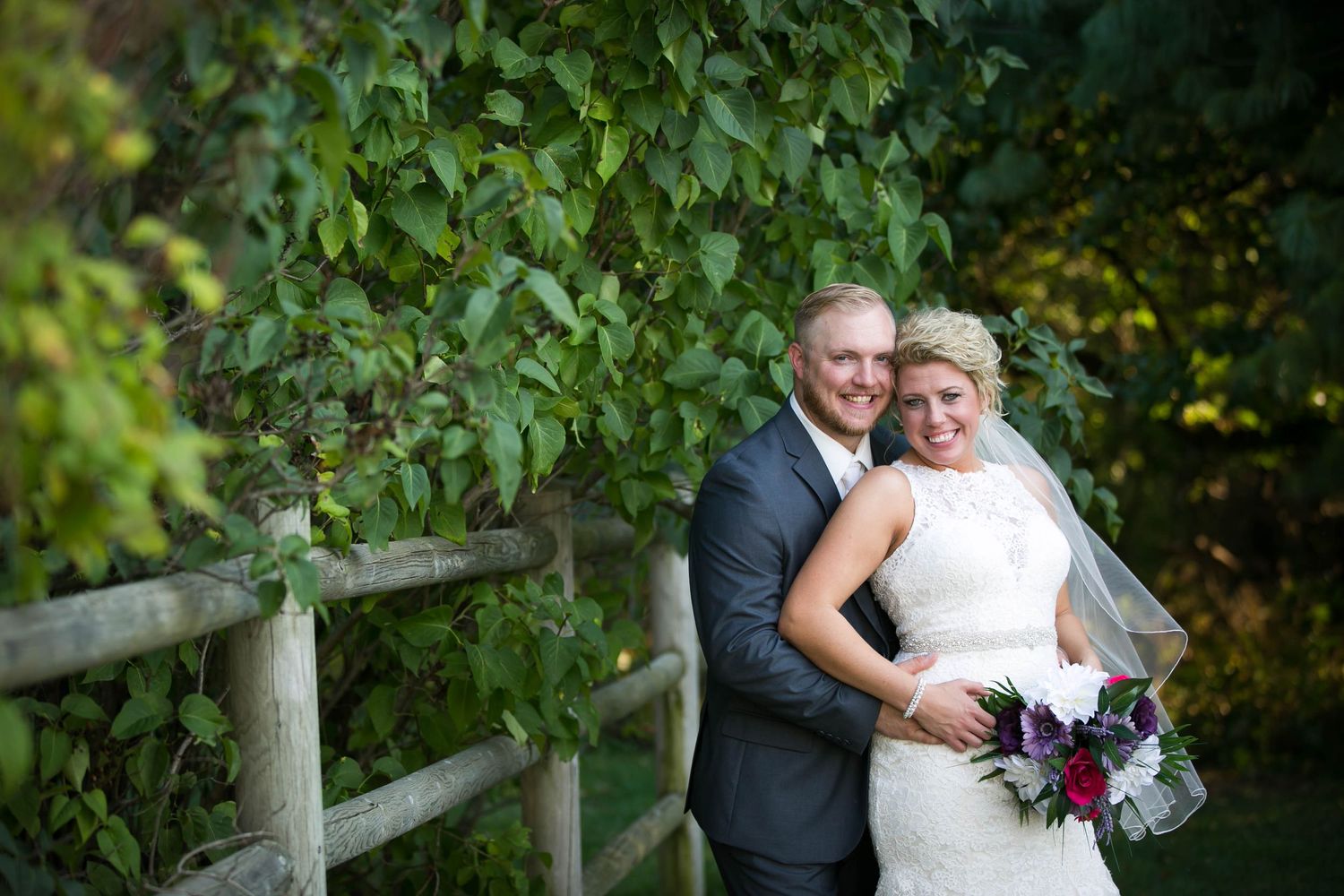 Bride and groom pose together near a rustic wooden fence surrounded by lush green foliage on their wedding day.