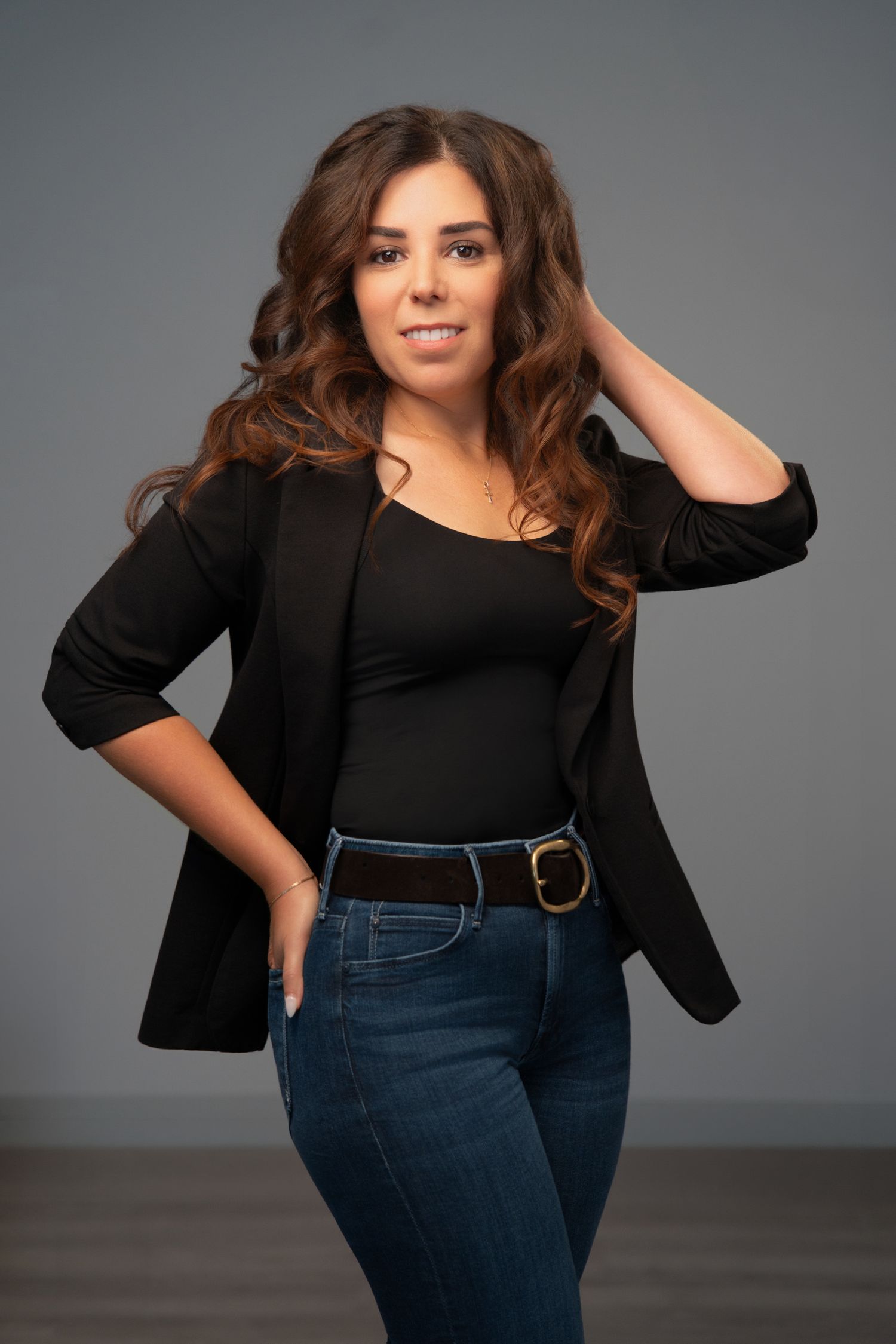Professional headshot of a smiling woman with curly brown hair wearing a black blazer against a gray background.