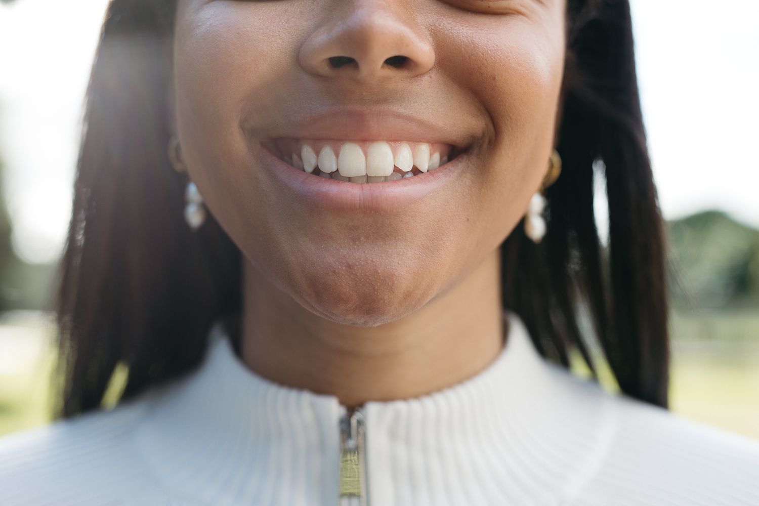 Close-up of a smiling young woman with white teeth, wearing pearl earrings and a white zip-up top outdoors.