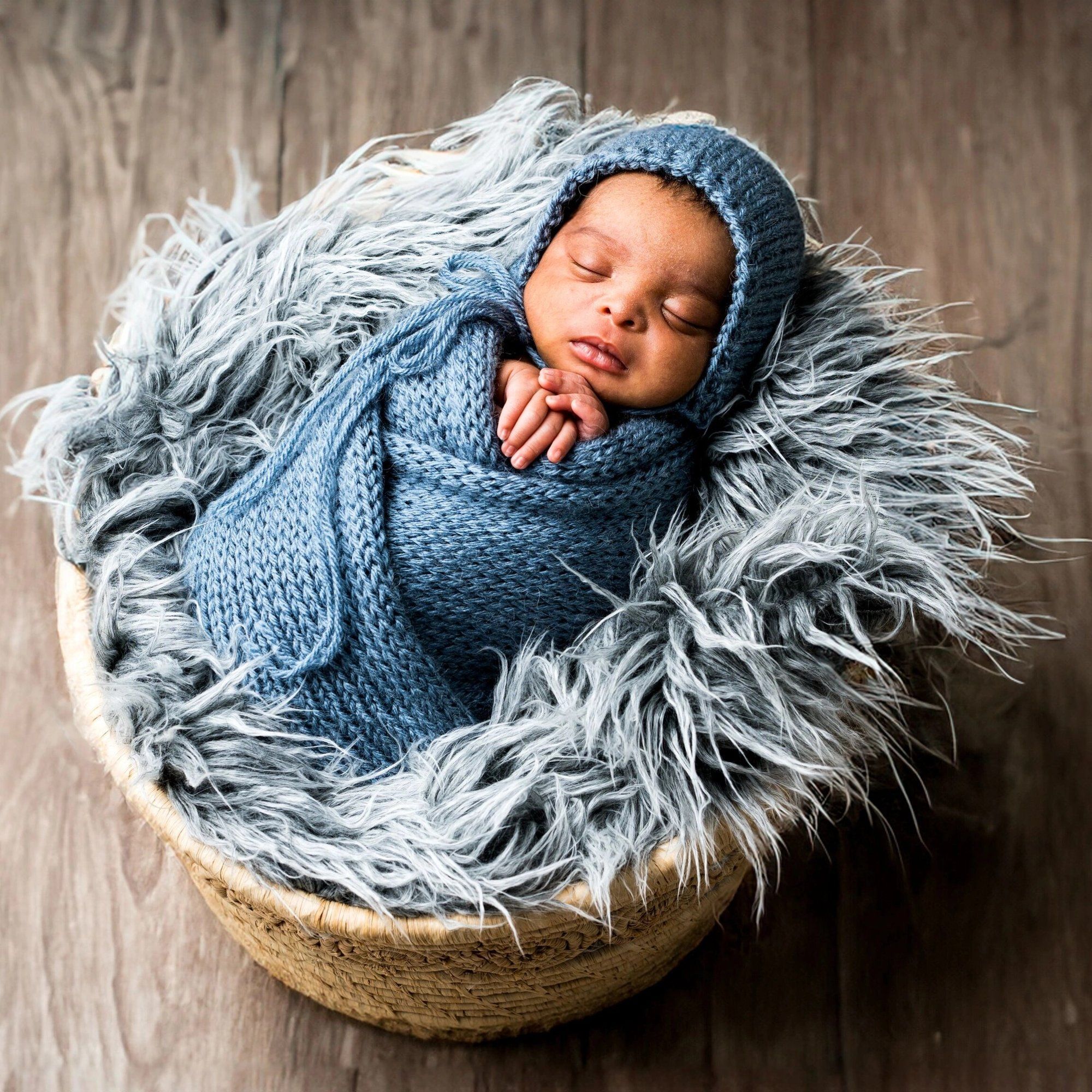 Sleeping newborn baby wrapped in blue knit blanket and bonnet, nestled in a wicker basket with fluffy gray fur.