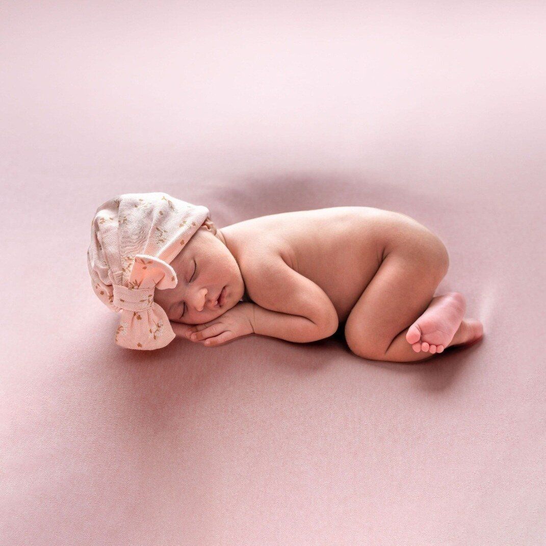 Peaceful newborn baby girl sleeping curled up on pink background wearing a delicate floral bow hat.