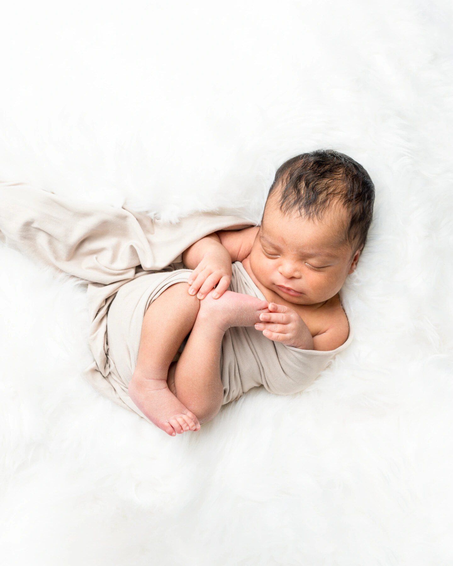 Sleeping newborn baby wrapped in a soft beige swaddle on a fluffy white fur blanket.