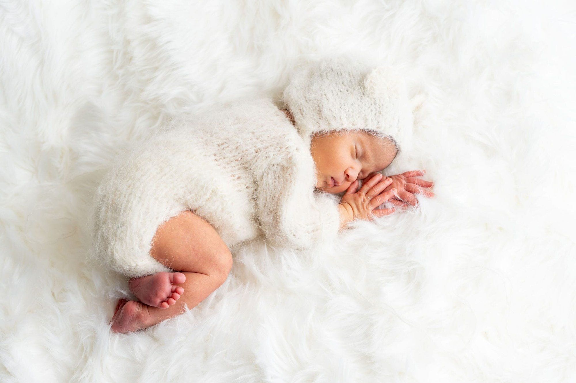 Newborn baby sleeping peacefully on white fur blanket wearing cozy knit white outfit and hat.