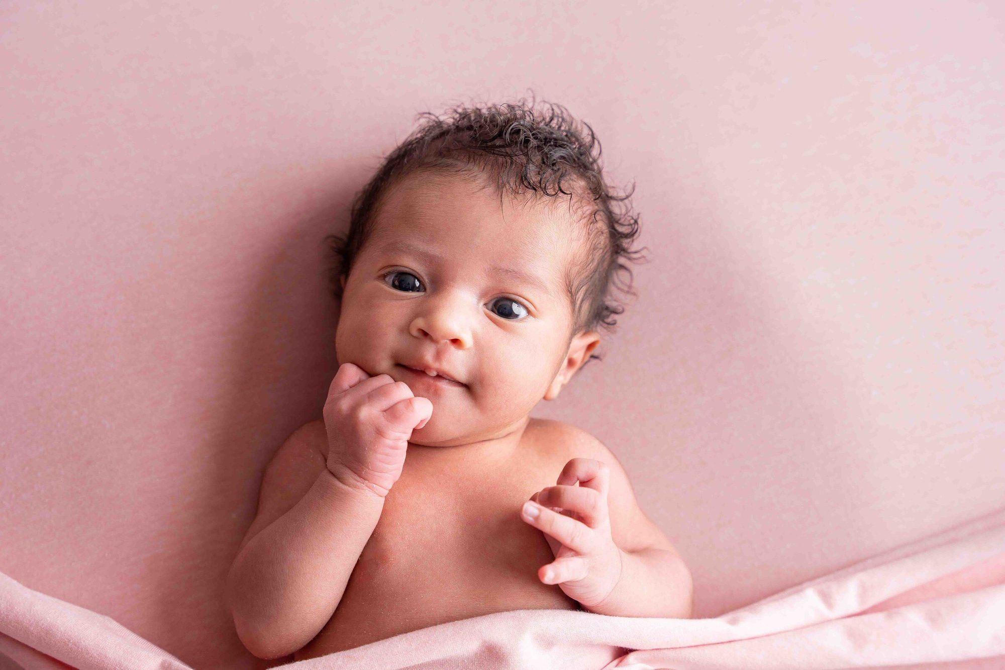 Newborn baby lying on pink blanket with hands near face, looking up with curious dark eyes.