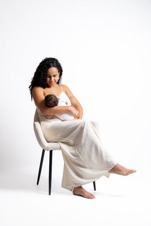 A mother sits on a wicker chair breastfeeding her newborn baby, wearing a white top and cream wide-leg pants.