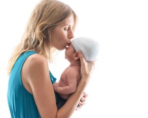 A mother in a teal dress tenderly kisses her newborn baby wearing a white hat against a white background.