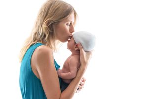 A loving mother in a teal dress kisses her newborn baby wearing a white hat against a white background.