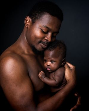 A shirtless father tenderly holds his newborn baby against his chest in a dark studio portrait.