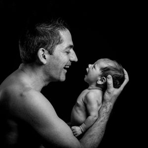 Black and white photo of a smiling father holding his newborn baby face-to-face against a dark background.