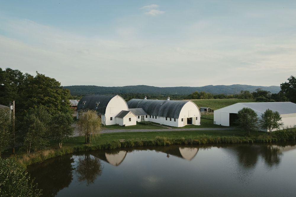 White barn buildings reflected in a calm pond with green trees and mountain backdrop under a partly cloudy sky.