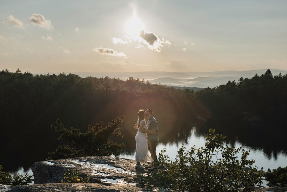 Couple embracing on rocky cliff at sunset with forest and lake views in a stunning outdoor wedding photo.