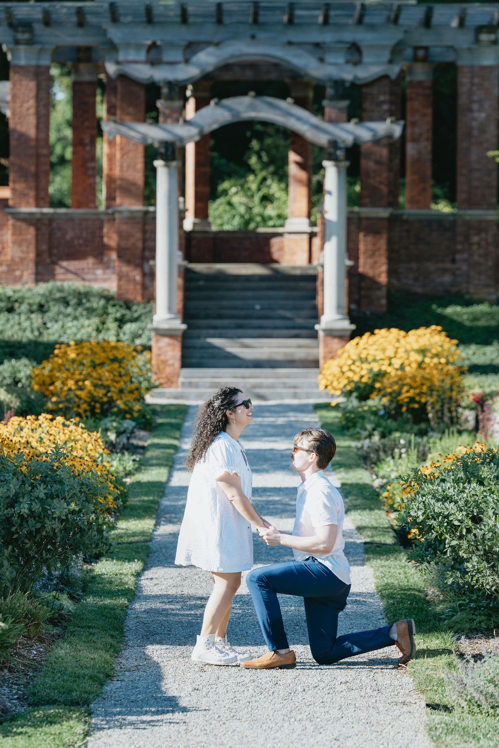 Man proposes on one knee to woman in white dress in a beautiful garden with brick archway and yellow flowers.
