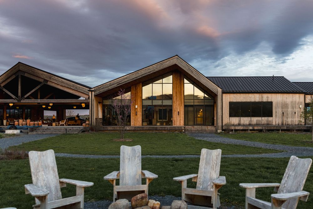 Rustic lodge with wood and metal architecture at dusk, featuring Adirondack chairs around a fire pit on a green lawn.