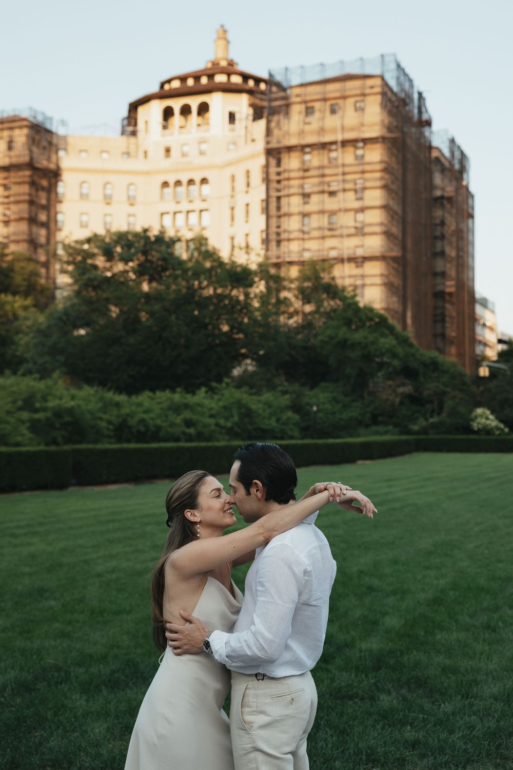 Couple embracing on green lawn with historic New York City building in golden hour light behind them.