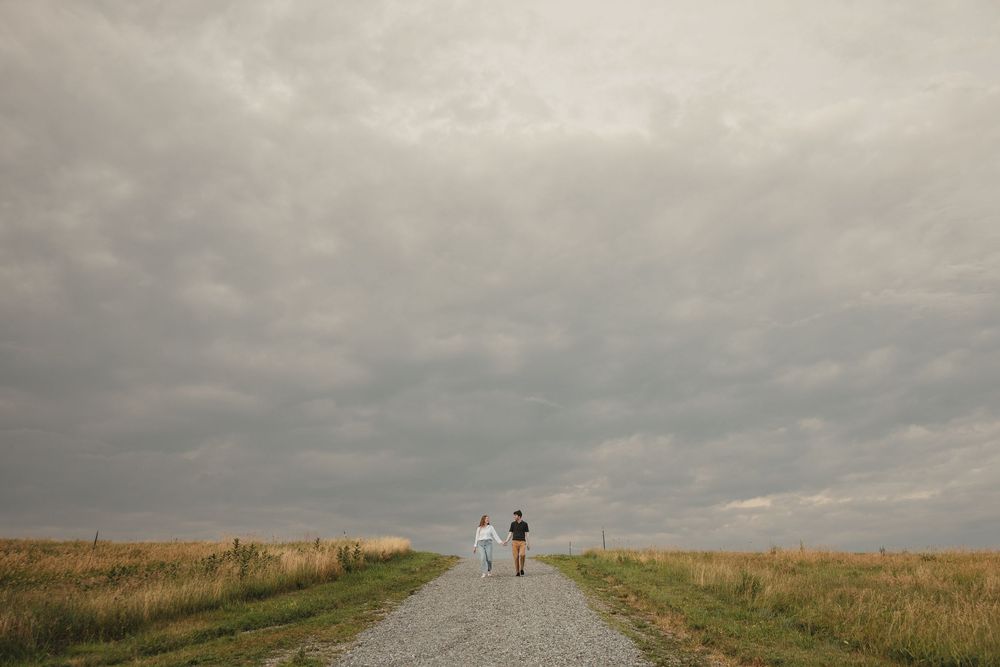 Couple walking hand in hand down a gravel path through open fields under dramatic cloudy skies.