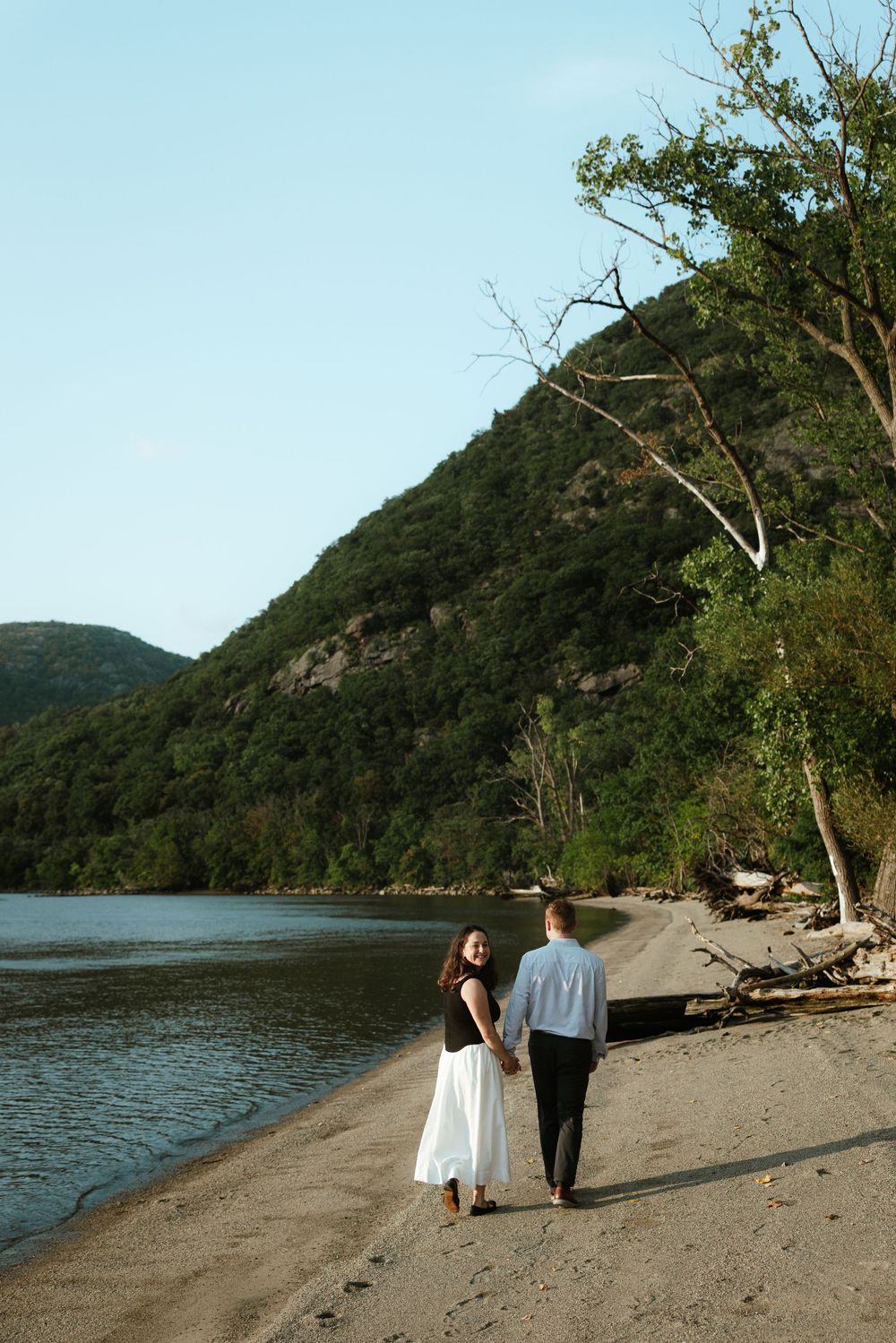 Couple holding hands walking along a sandy riverbank with lush green mountains in the background on a clear day.