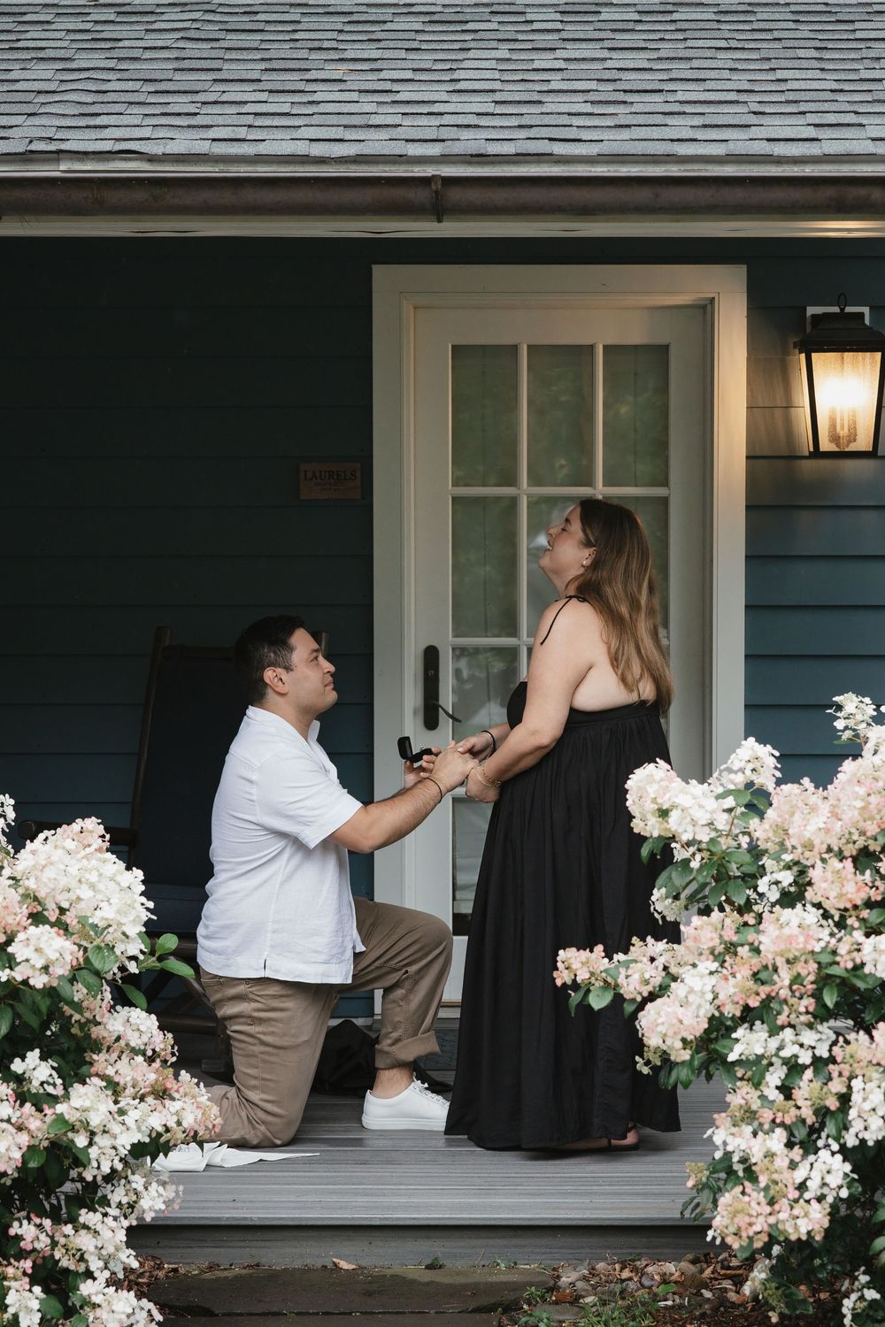 Man kneeling on porch proposing to woman in black dress, surrounded by white hydrangeas at blue house entrance.