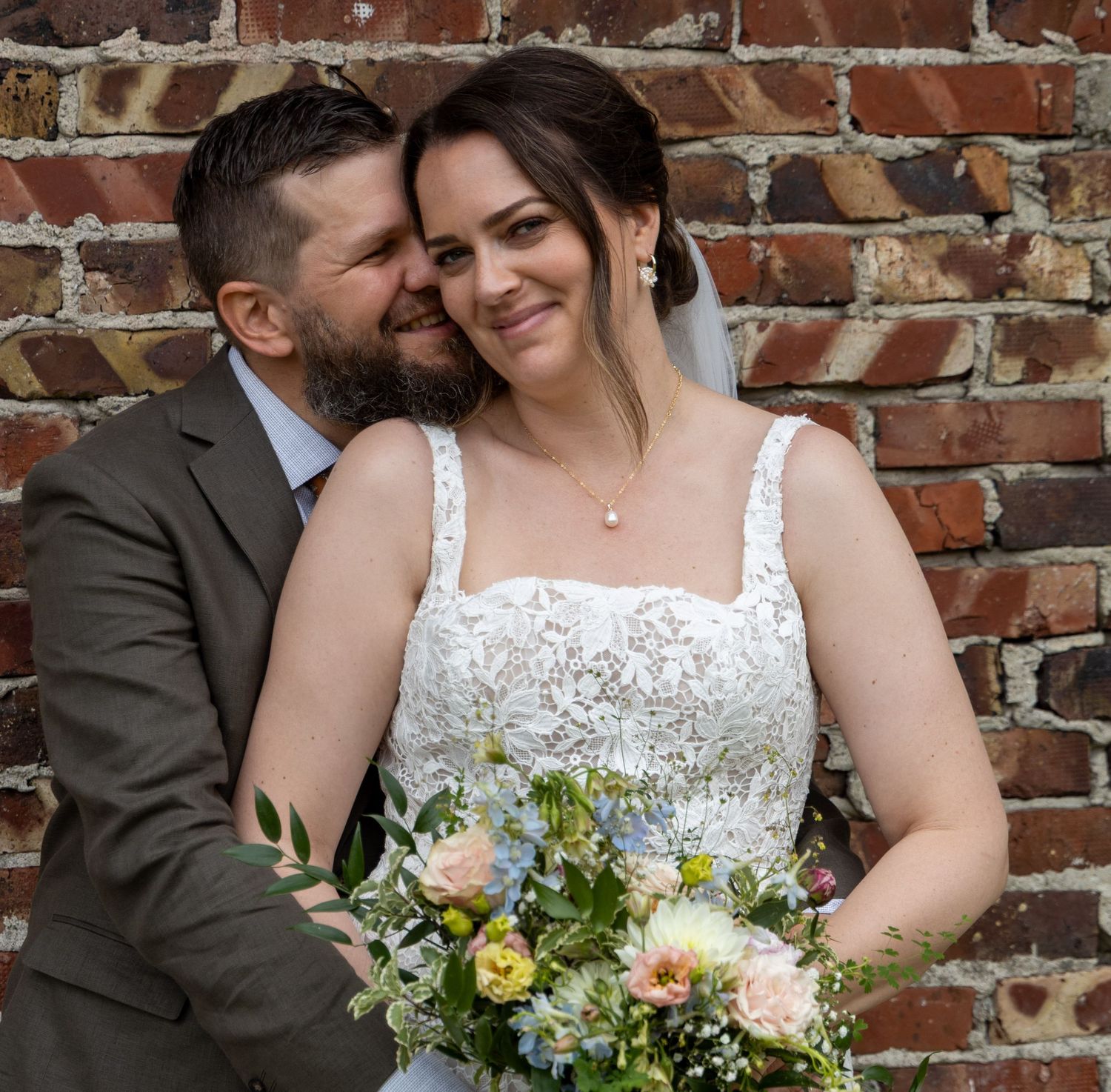 A bride and groom pose romantically against a brick wall, she holds a pastel bouquet in a lace gown.