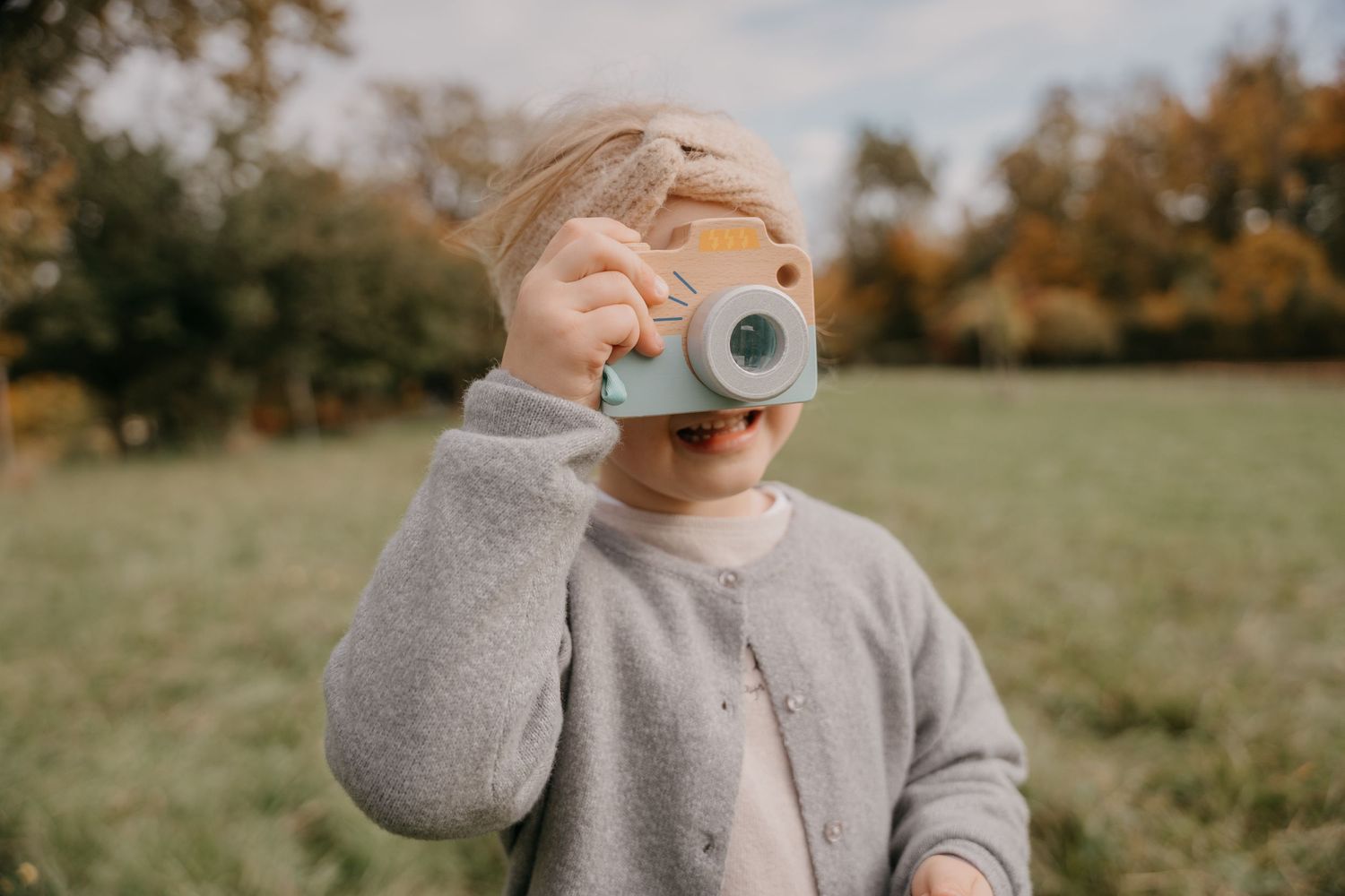 A young child holds a toy camera up to their face outdoors in a green field with autumn trees in the background.