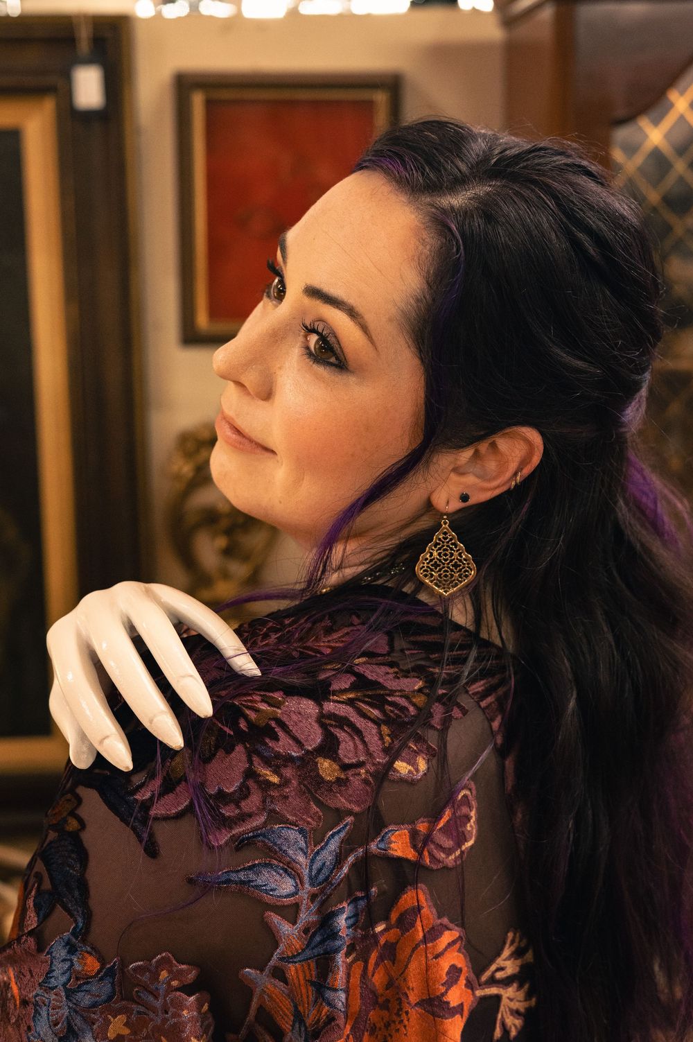Dark-haired woman in floral embroidered top with gold earring glances over shoulder in art gallery.